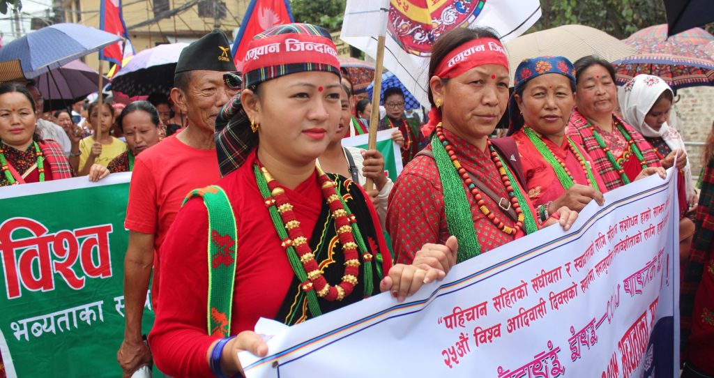Image focuses on four people, dressed in traditional outfits, holding a banner with group of people following them fading out in the background.